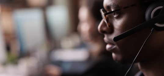 Helpdesk secretary listening to client on helpline call, using customer service network on headset. Male receptionist working at call center telemarketing assistance. Close up.